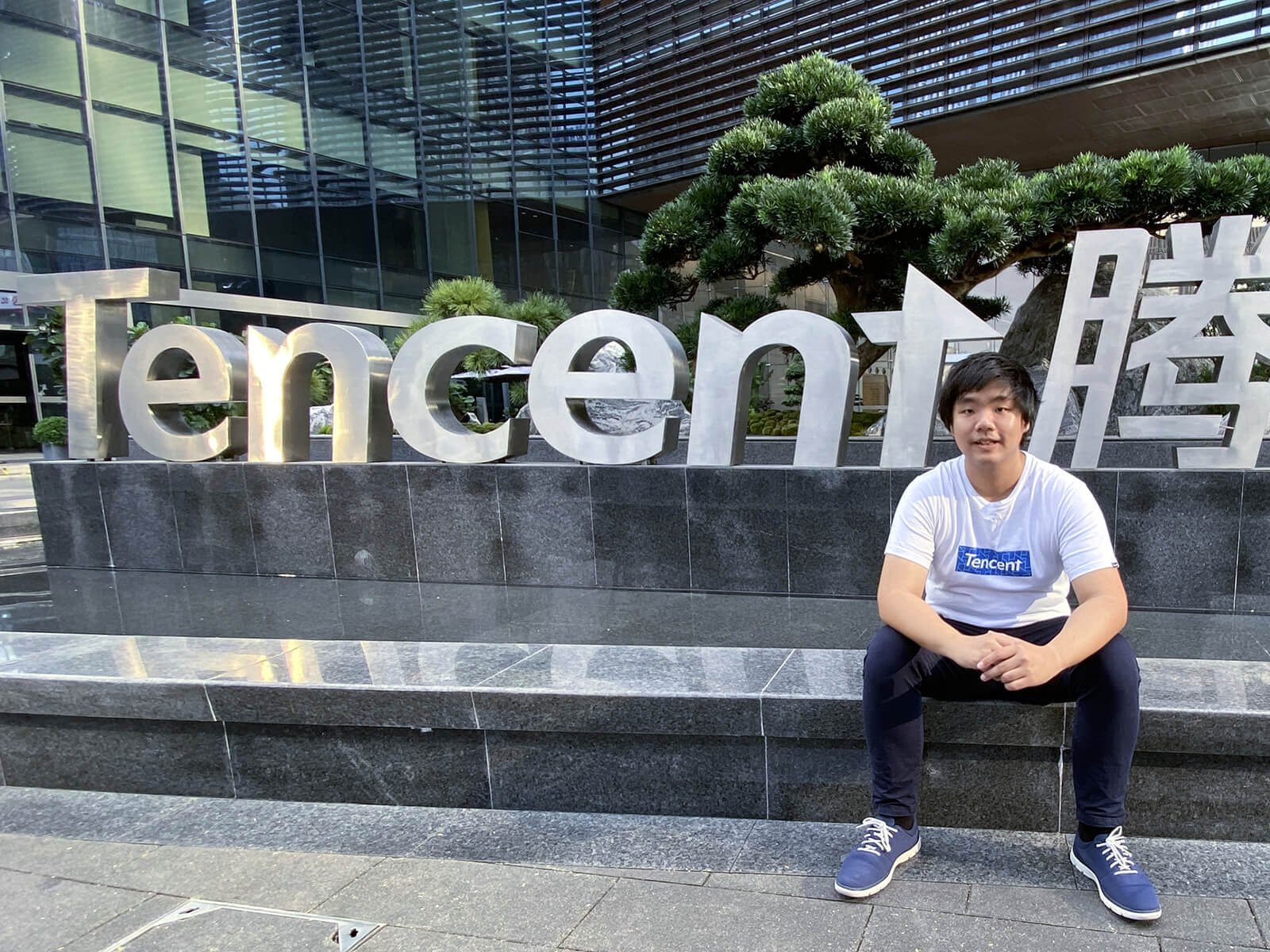 Sim Yi Cong sits by a pool in front of a large Tencent logo sign outside the Tencent headquarters building.