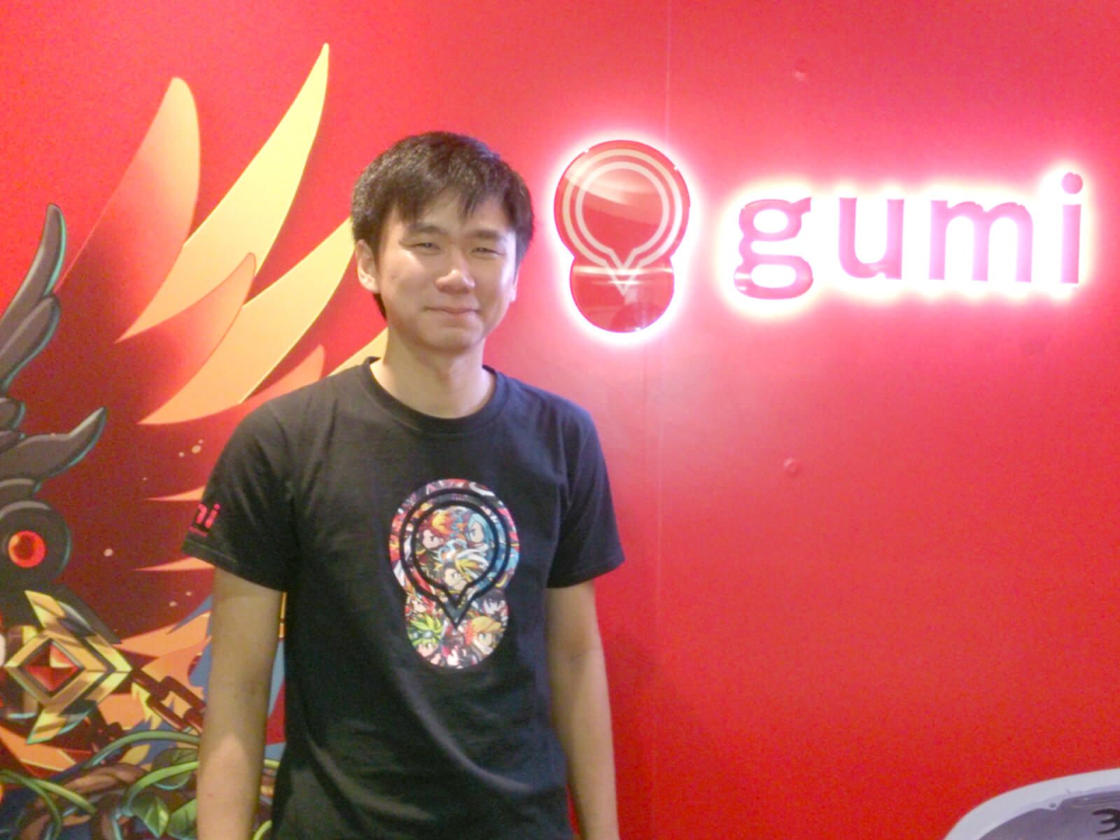 DigiPen graduate Soh Huajin stands in front of a red wall with a glowing logo for gumi Asia behind him