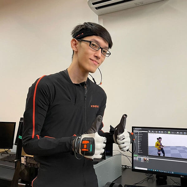 Tay How Wen wears mocap hardware with two thumbs up next to a computer monitor