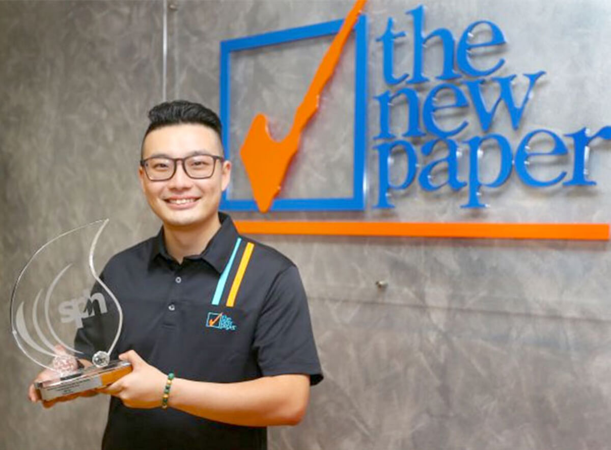 DigiPen graduate Billy Ker holds a glass droplet shaped award reading "SPH" in front of a wall with The New Paper's logo