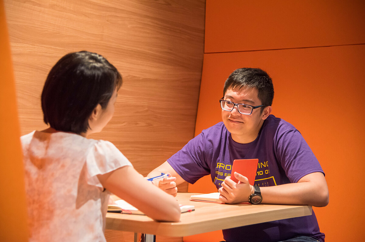 Chester Liew sits at a table in an orange-colored alcove, talking to a co-worker