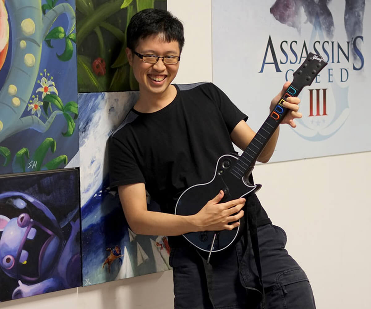 Chin Jia Hao poses smiling holding a black Guitar Hero peripheral. Posters for Assassin's Creed and Rabbids are behind him