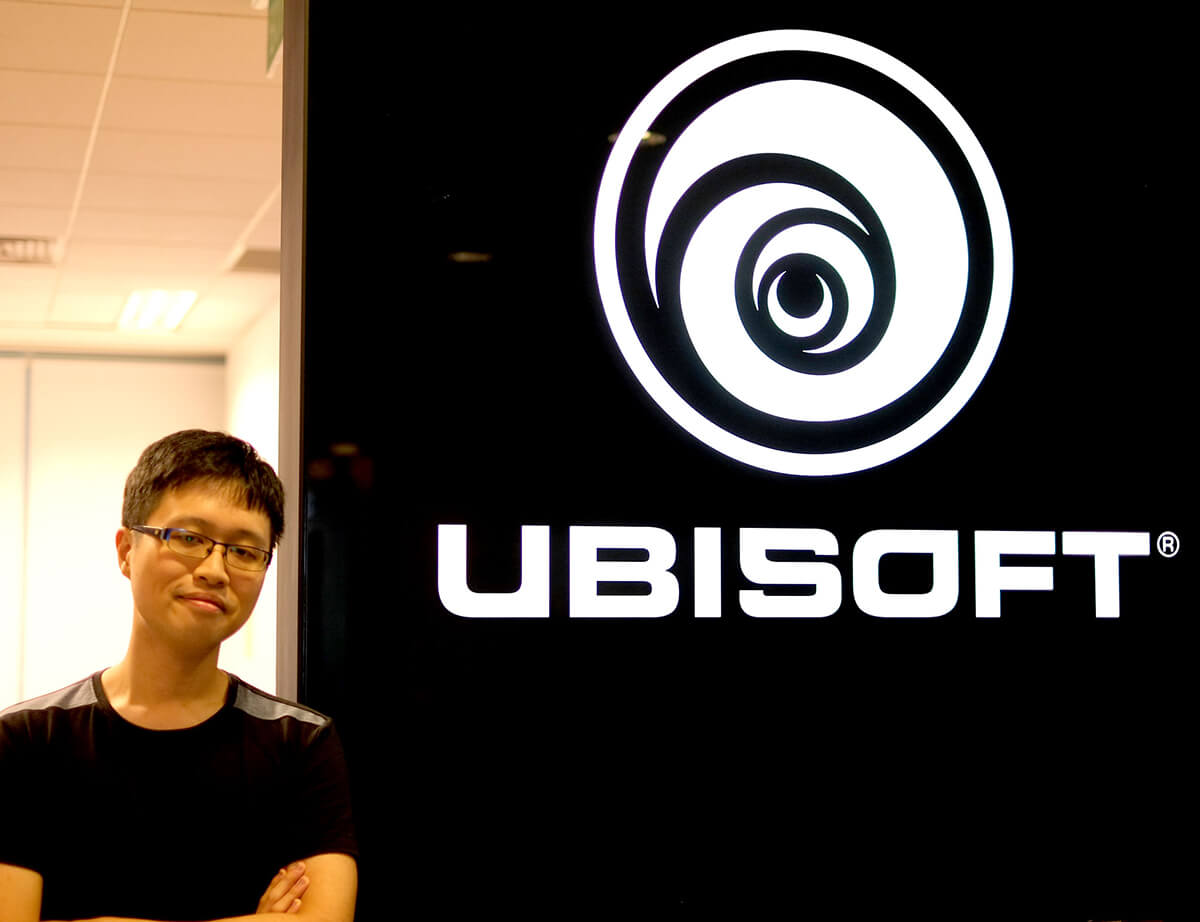DigiPen graduate Chin Jia Hao stands with arms crossed next to black-and-white Ubisoft logo