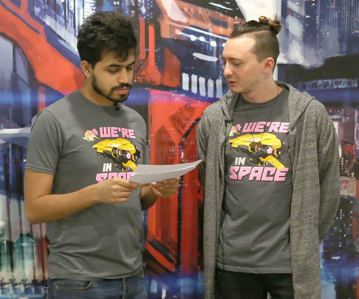 Two members of Daylight Studios stand in gray t-shirts reading "We're in Space" looking at a piece of paper