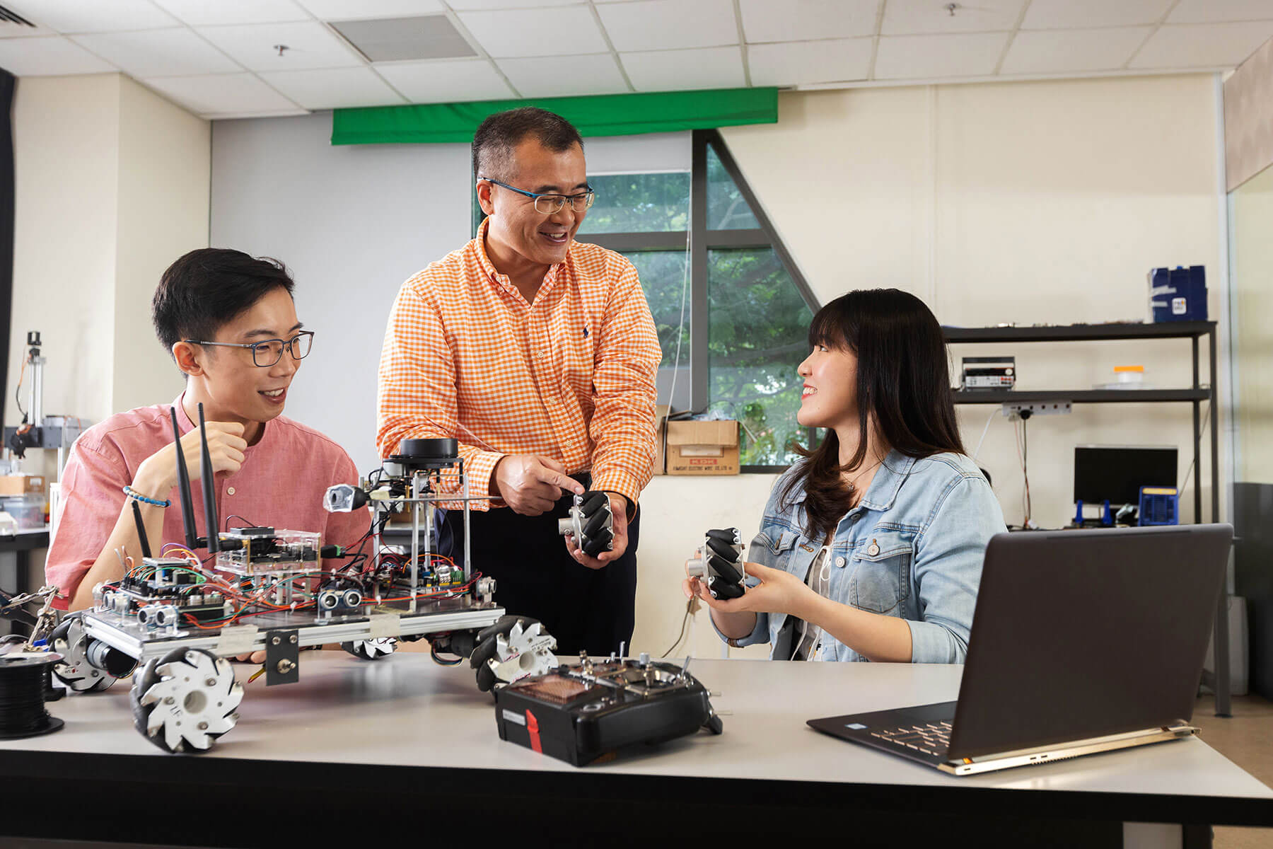 Dr. Tang Liang stands next to two students while explaining a device in his hand to them.