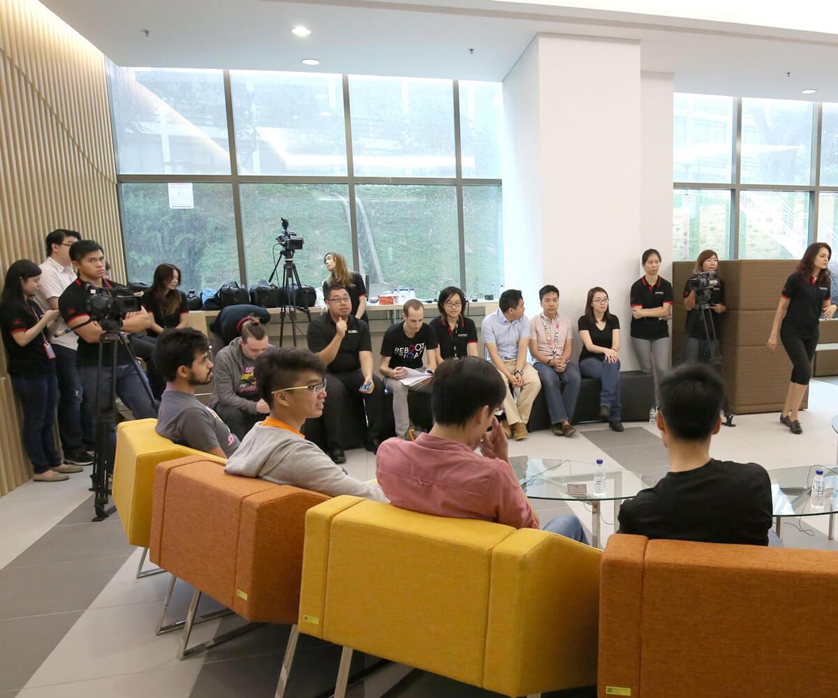 Game industry professionals from behind seated in orange and yellow chairs in a foyer being filmed by cameras