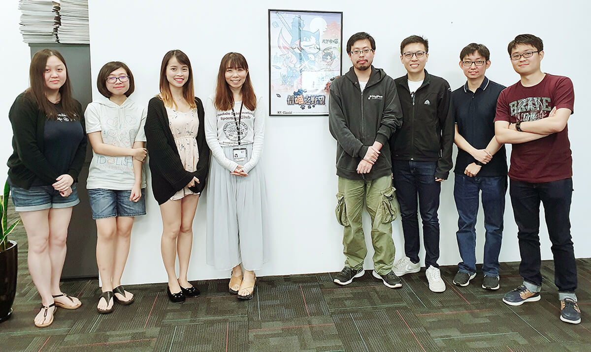 Group photo of the 8 DigiPen (Singapore) alumni who worked on Koei Tecmo's Nyapuri standing in office by a poster of the game.