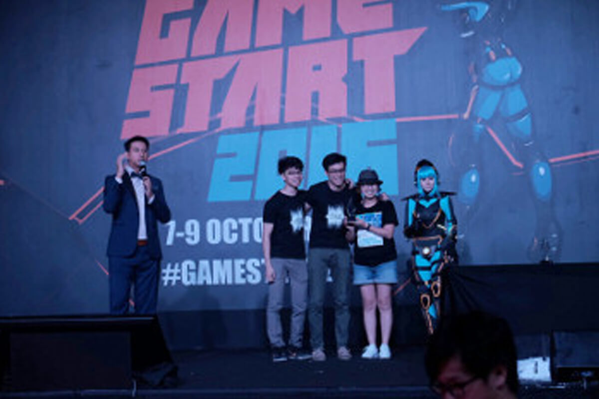 Three members of the development team standing onstage with emcee and cosplayer. "Game Start 2016" on a banner behind them.