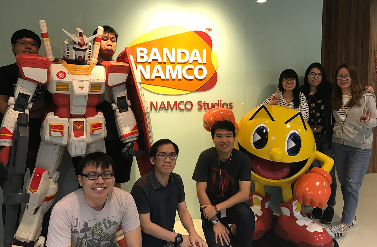 Group photo of eight DigiPen alumni in the lobby of Bandai Namco studio, posing by statues of Pac-Man and a mecha robot.