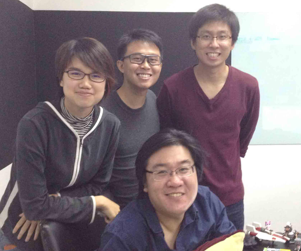 Three graduates stand posing with Creative Director Ian Gregory Tan seated at a desk