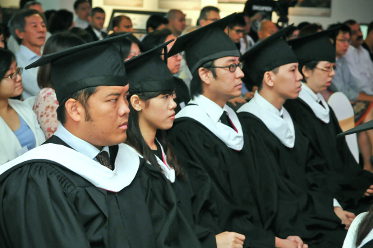 Five seated students watch the graduation ceremony in their cap and gowns