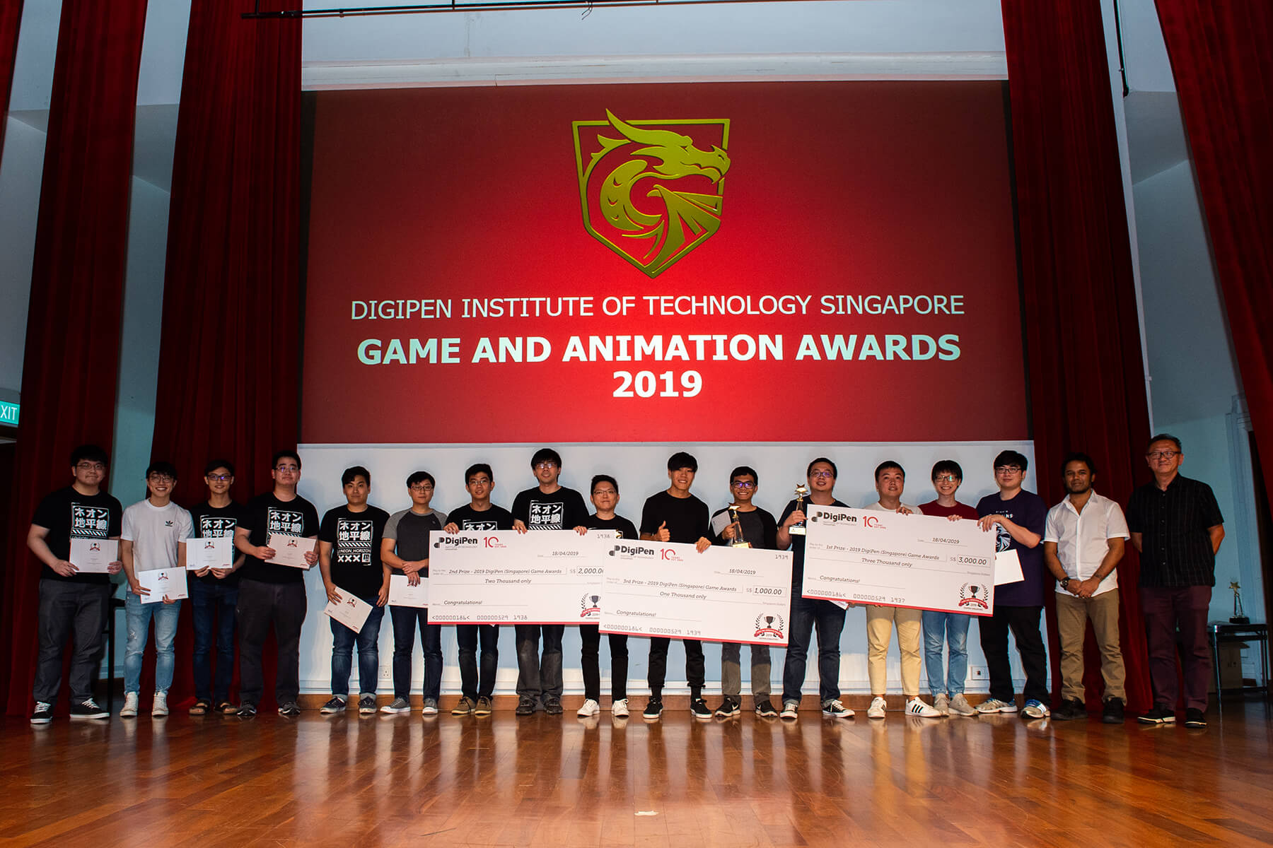 16 students line up in a row to show off their giant checks, certificates, and trophies during the DigiPen Game and Animation Awards show.