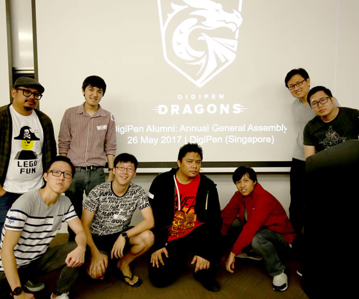 Members of the alumni committee stand in front of a digital projection screen with the DigiPen Dragon logo on it