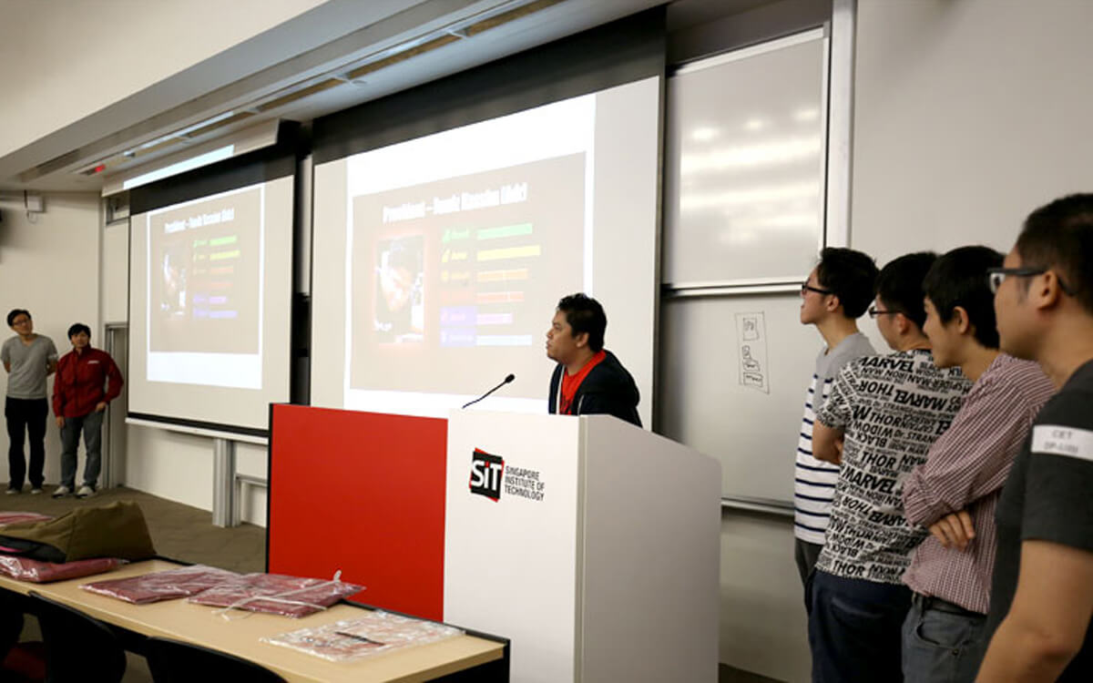 A member of the alumni committee stands at a lectern next to digital projection screens during a speech