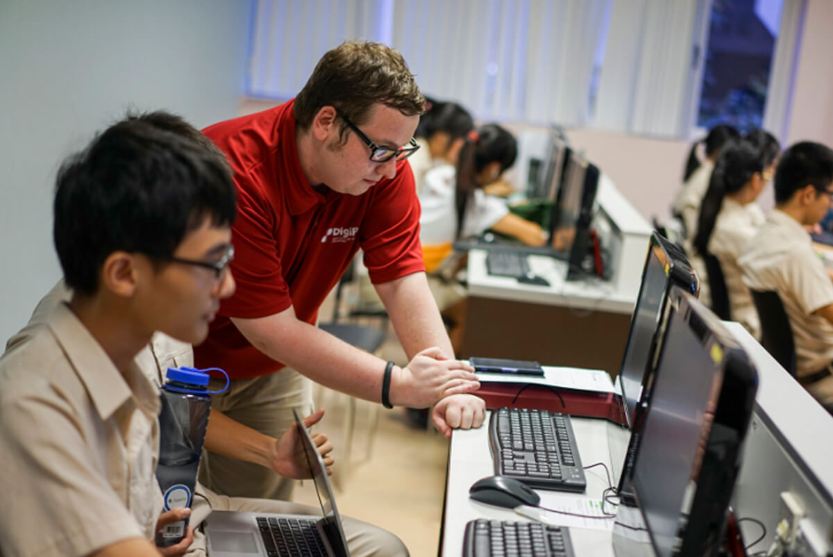 An instructor from DigiPen (Singapore) in a red shirt leans over at a desk looking at a student's computer monitor