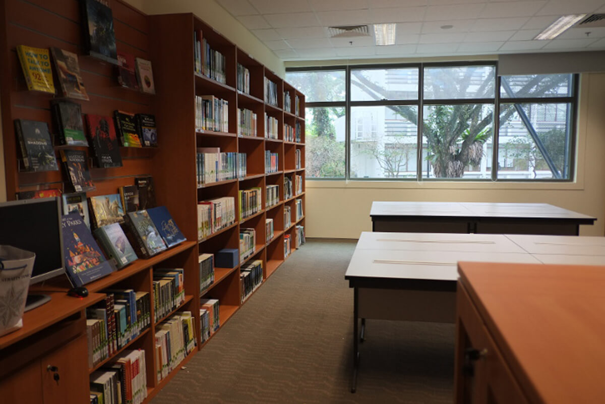 Library area with left wall taken up by wooden bookcases full of books. Windows in the far wall, and desks in the center.