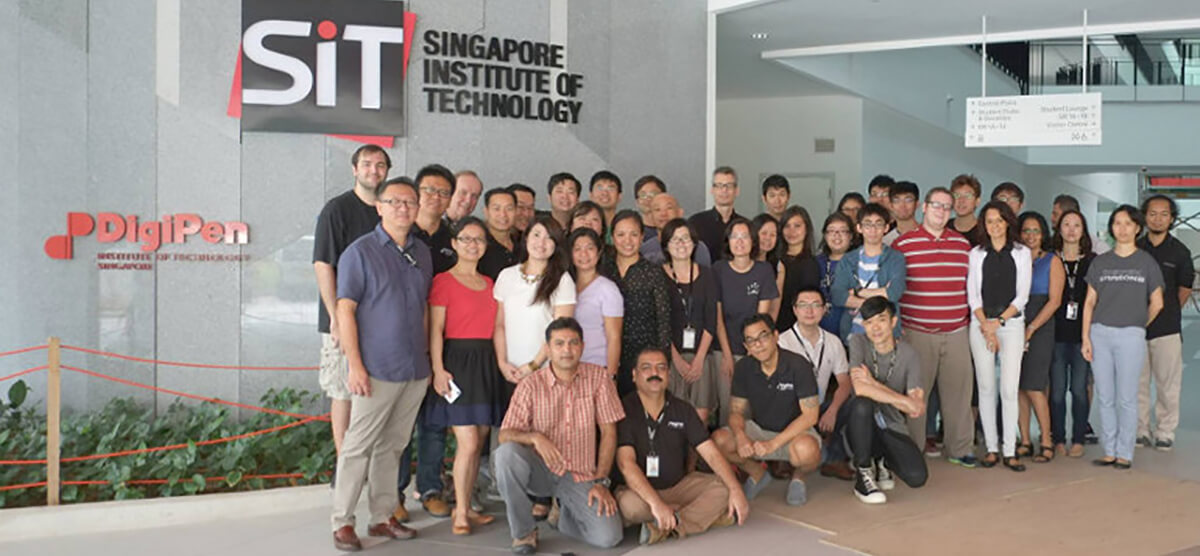 Dozens of DigiPen staff pose for a photo in front of a gray marble wall with the DigiPen and SIT logos