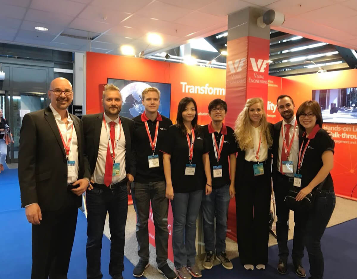 Group photo of 8 Visual Engineering employees standing together and facing camera in front of a company booth during an expo.