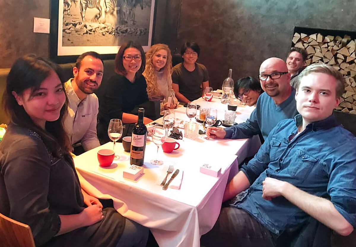 Photo of 9 Visual Engineering employees gathered around a restaurant table with white tablecloth and smiling toward the camera.