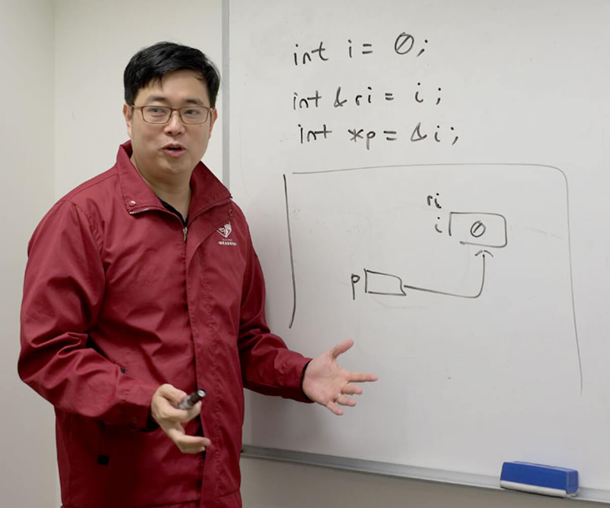 Dr. Edward Sim in a red DigiPen Dragon jacket explaining a diagram on a whiteboard to his class