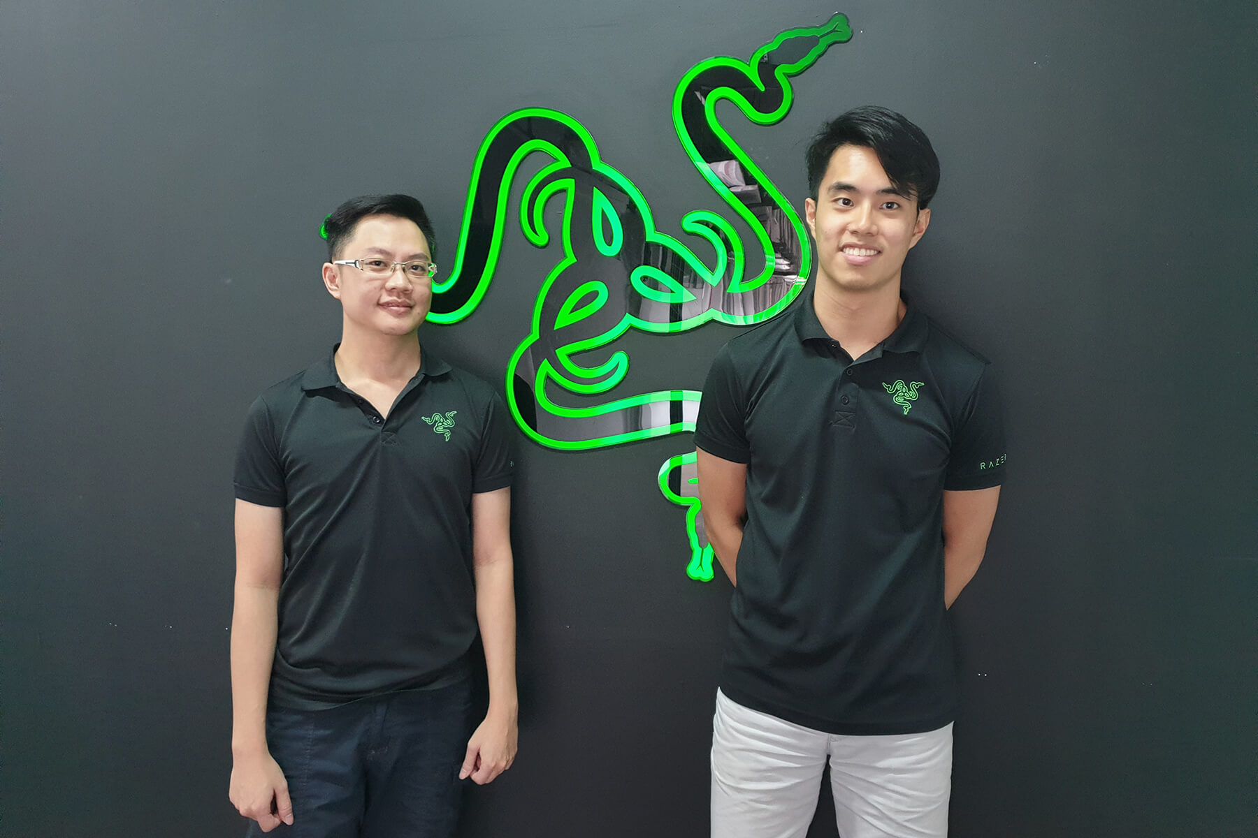 Two men wearing polo shirts with the green Razer logo stand by a gray wall, on either side of a much larger Razer logo.