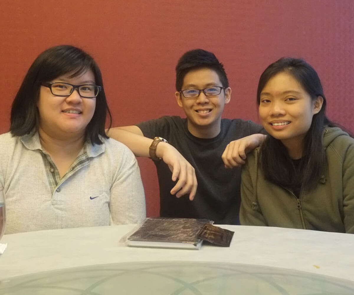 Graduates Gina Chiang, Trey Chua, and Jasmine Cheah pose for a photo sitting at a table in front of a red wall