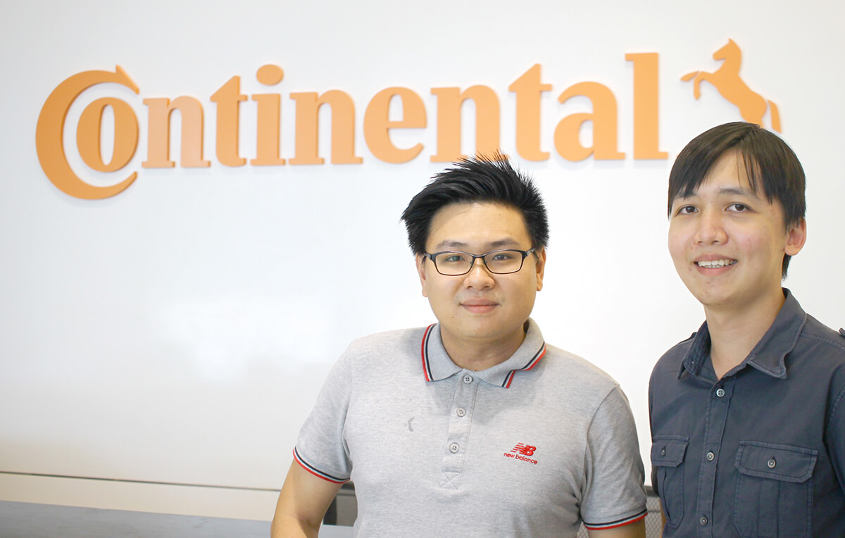 DigiPen (Singapore) alumni Jason Ngai and David Seah pose for a photo in front of a white wall adorned with the orange Continental logo