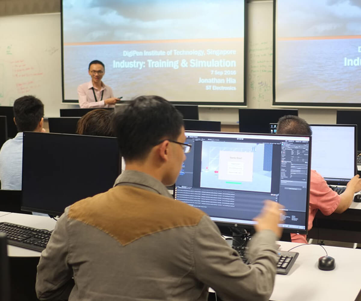 A student working at a computer while Jonathan Hia gives a presentation at the front of a classroom