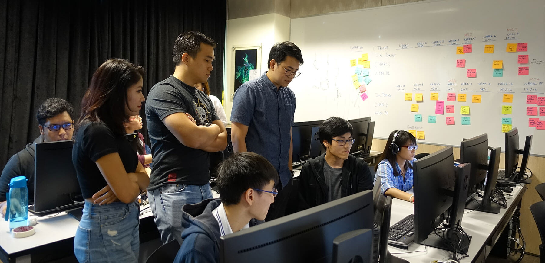 Kobe Sek from Ubisoft Singapore and others look down at a student’s monitor in a computer lab classroom, with students seated at two rows of workstations.