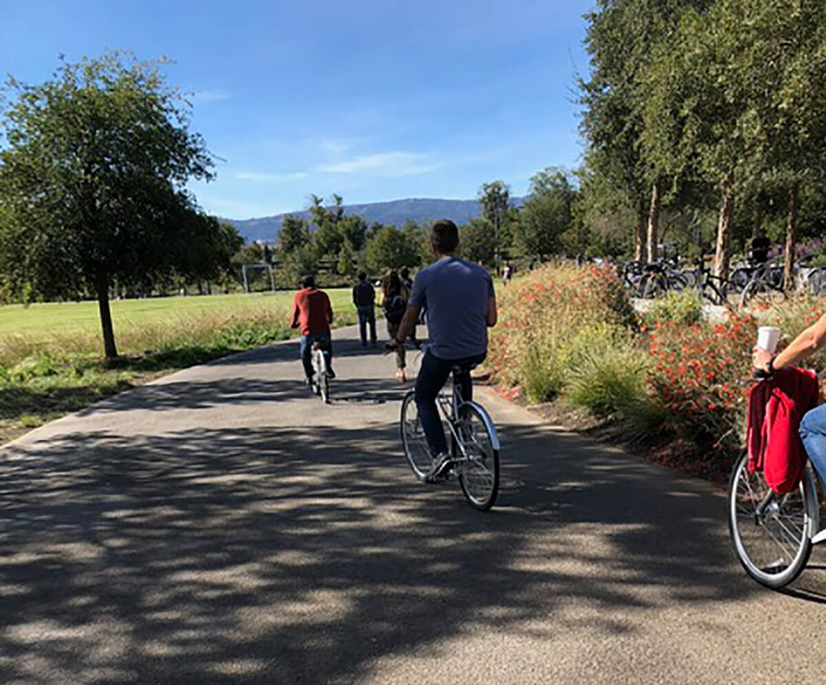 Photo of people riding bikes and walking under a clear blue sky along a paved outdoor pathway.