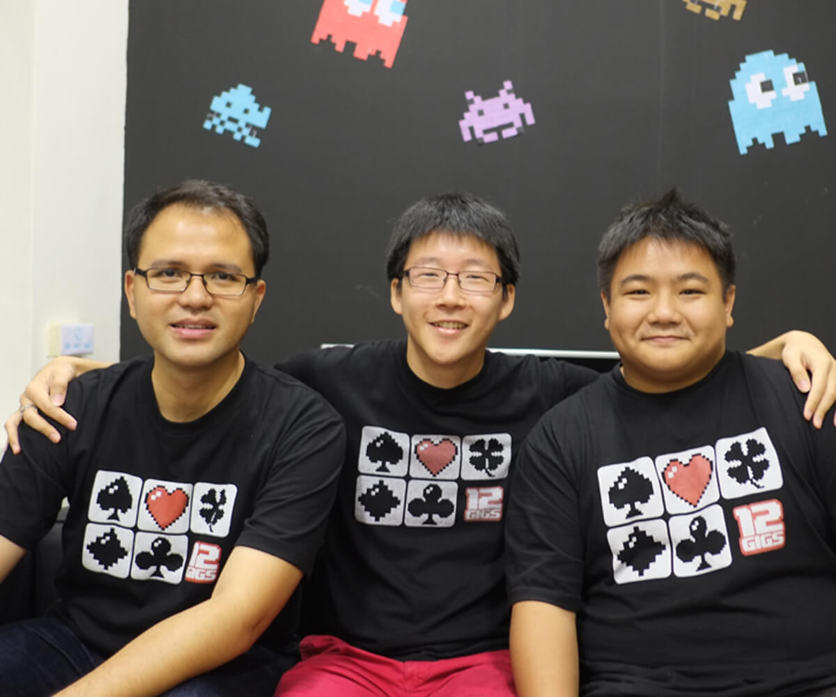 Three DigiPen graduates sit together in front of a wall decorated with pixel art and the 12 Gigs Logo wearing black t-shirts