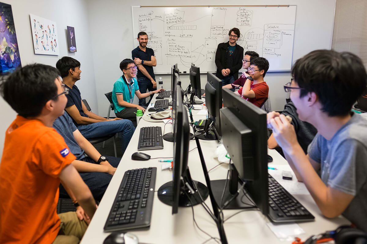 Several students sit at a long desk laughing as do their instructors, standing at a white board at the end of the table