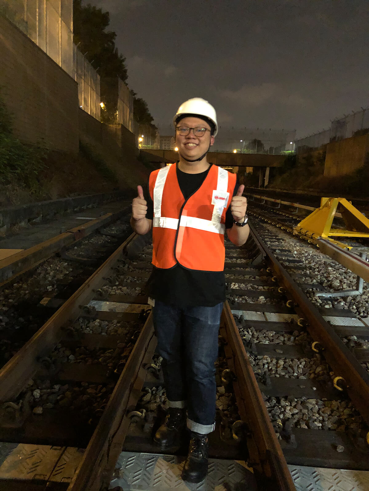 Samuel Tan dons a worker's vest and gives two thumbs up while standing on train tracks at night.