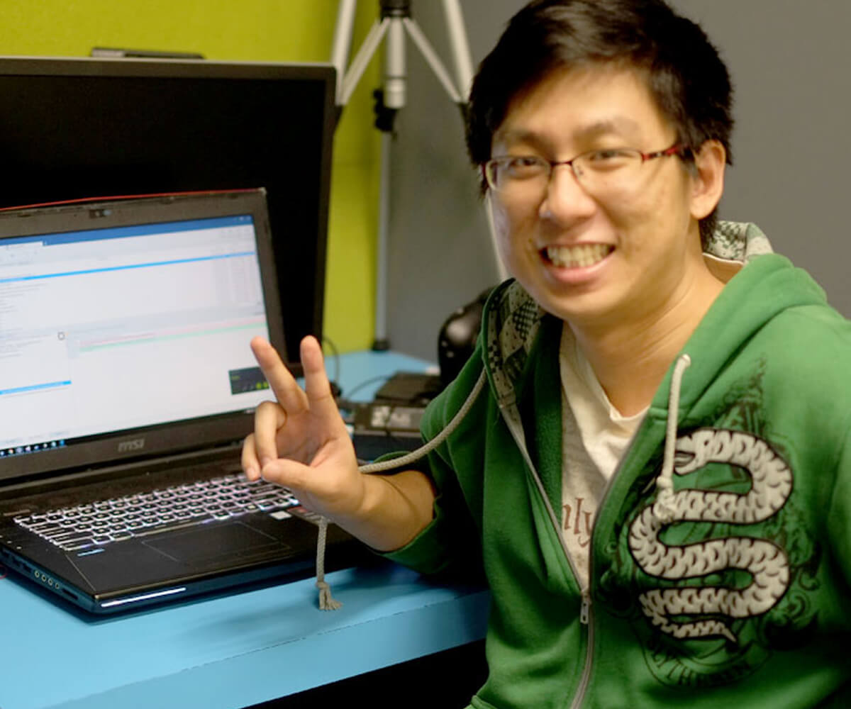 DigiPen graduate Sim Wei Jin seated at a desk with a laptop posing for a photo while giving a V sign with his hand