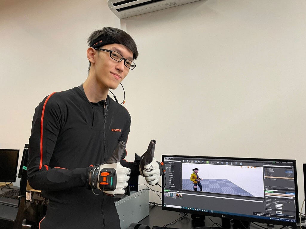 Tay How Wen wears mocap hardware with two thumbs up next to a computer monitor