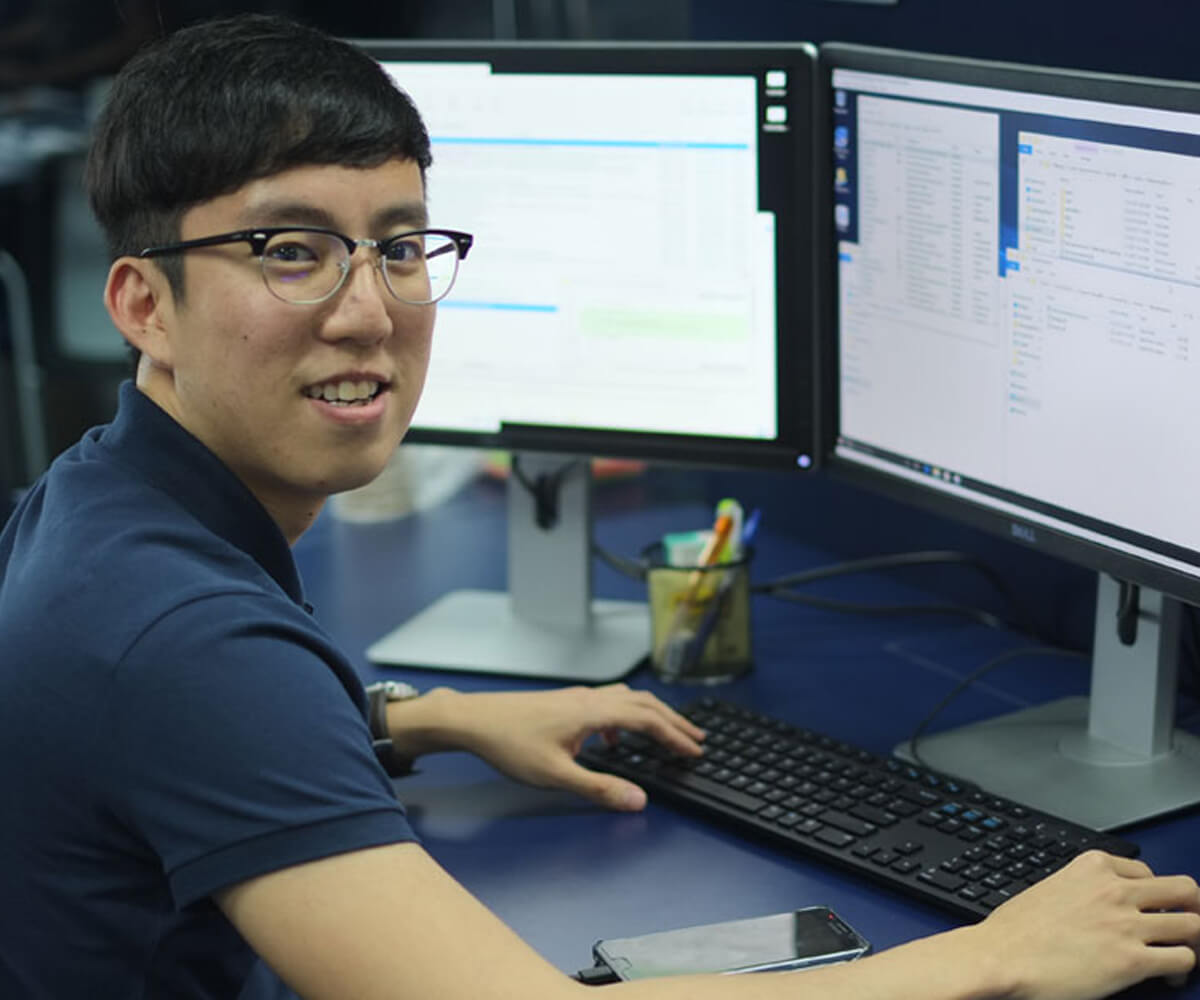 DigiPen graduate Woon Kok Leong poses for a photo seated at a computer on a blue desk